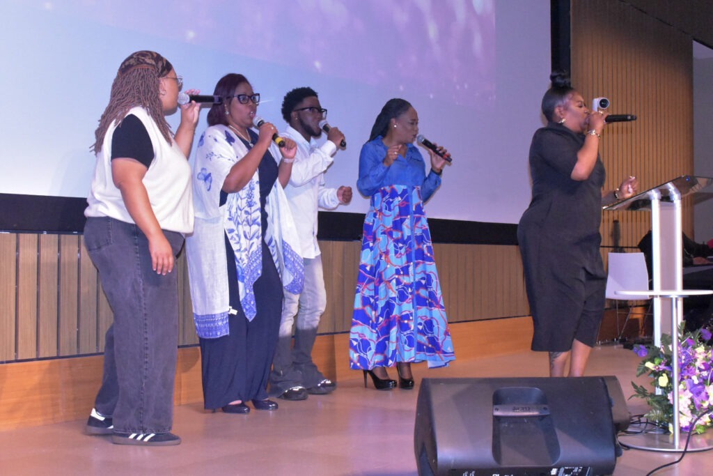 Five vocalists perform on stage with microphones, showcasing a lively music event. The backdrop features soft lighting, enhancing the vibrant atmosphere.