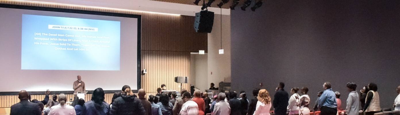 A congregation worships during a service, facing a screen displaying "Righteous and Glorious, Ever Victorious." The atmosphere is vibrant and uplifting.
